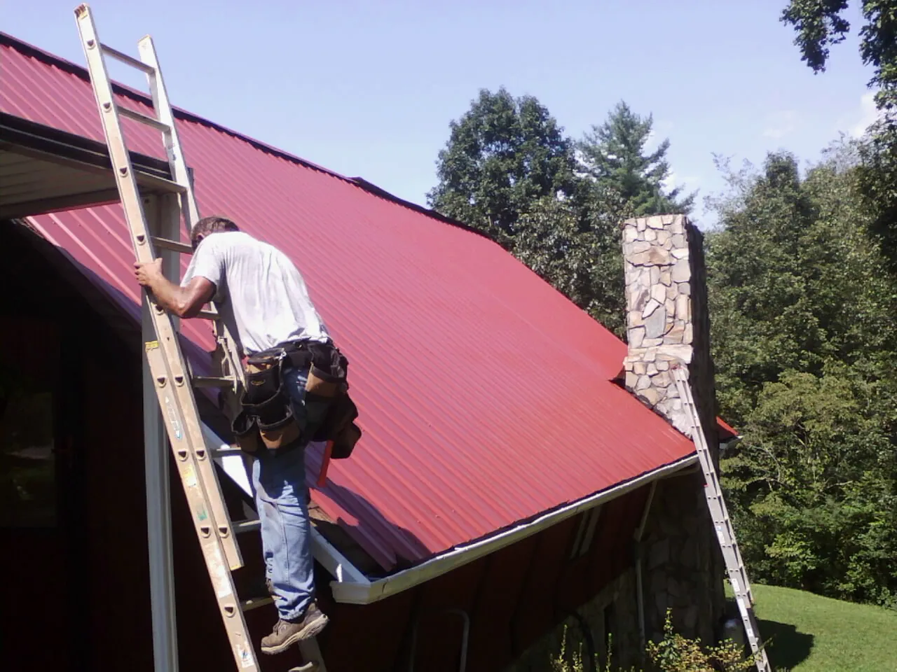 Roofer on a ladder beside a red metal roof and stone chimney — Soffit & Fascia Repair by Roofing Brothers in Reubens